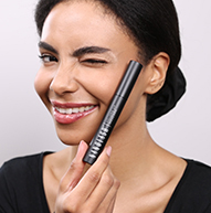 A woman smiles brightly while holding a mascara brush
